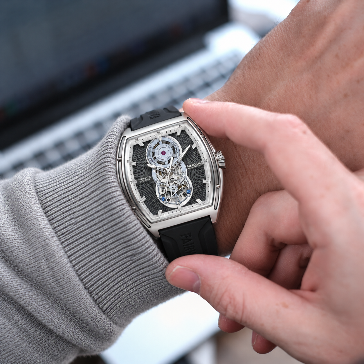A close-up lifestyle shot of a men's skeleton watch with a black rubber strap and silver-toned case, worn on a wrist and being adjusted by another hand in front of a blurred laptop background.