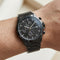 Black wristwatch with detailed face on a wrist against a neutral background