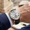 Elegant automatic skeleton watch on wrist with white Roman numeral dial, visible tourbillon, black leather strap, and tailored suit sleeve in warm natural light