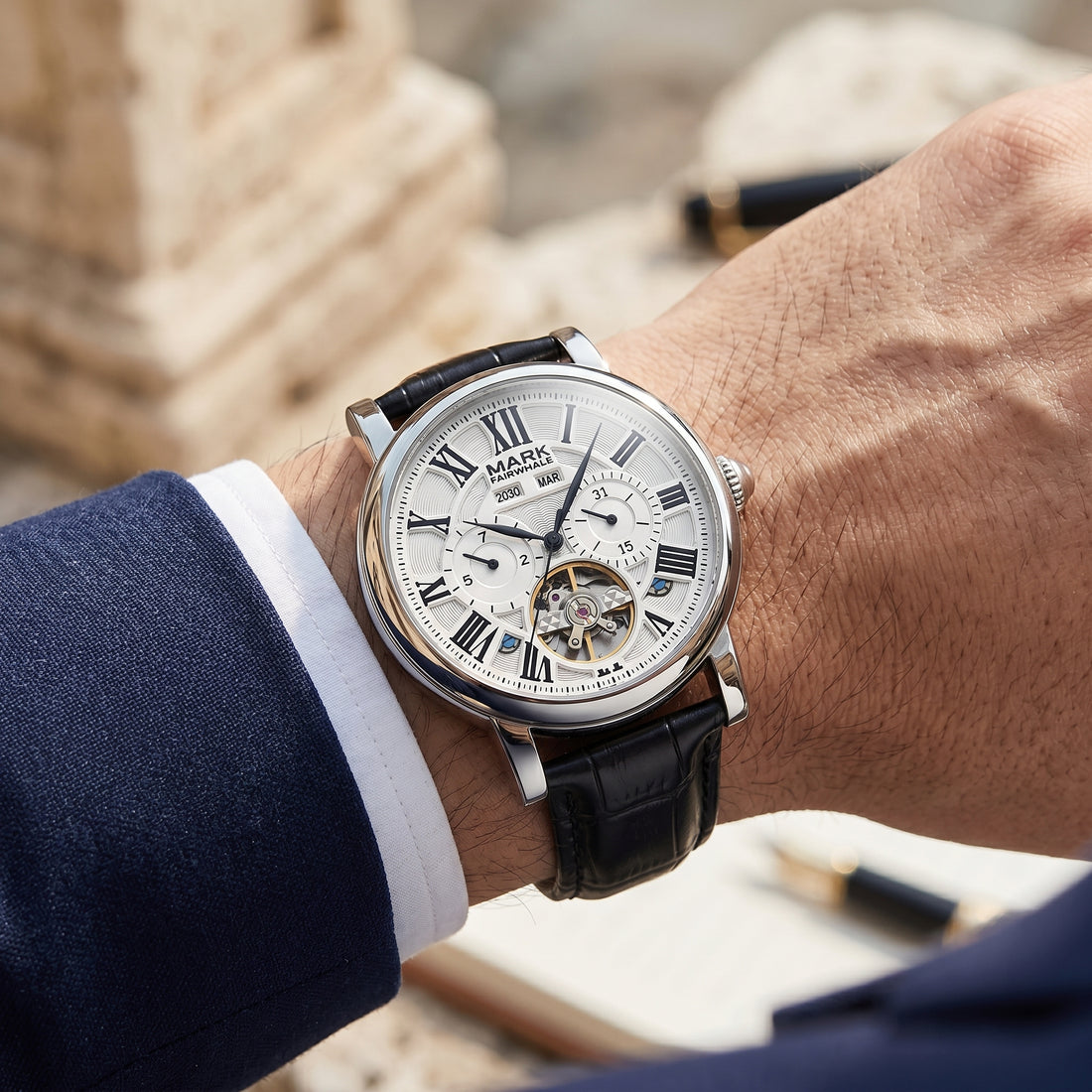 Elegant automatic skeleton watch on wrist with white Roman numeral dial, visible tourbillon, black leather strap, and tailored suit sleeve in warm natural light