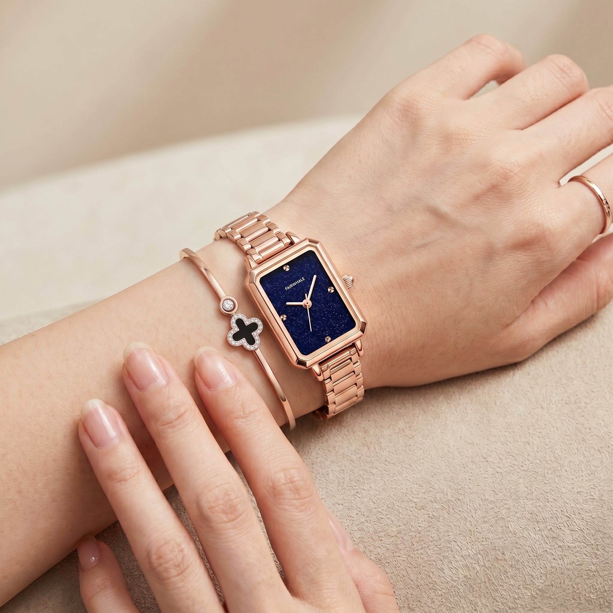 Elegant rose gold rectangular watch on woman wrist with deep blue dial, minimalist markers, and matching bracelet jewelry in soft neutral setting