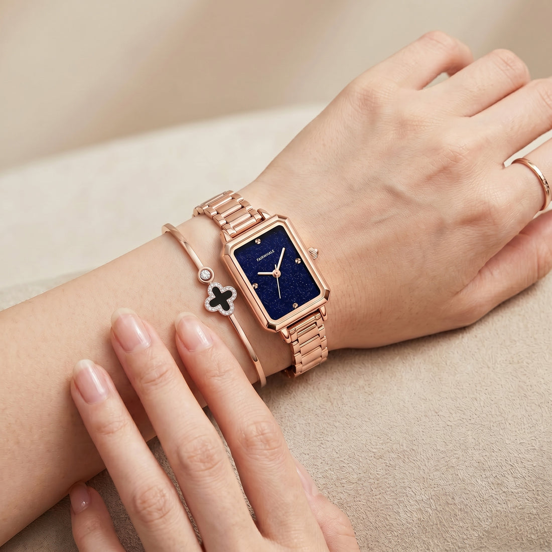 Elegant rose gold rectangular watch on woman wrist with deep blue dial, minimalist markers, and matching bracelet jewelry in soft neutral setting