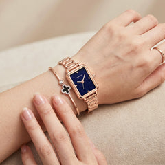 Elegant rose gold rectangular watch on woman wrist with deep blue dial, minimalist markers, and matching bracelet jewelry in soft neutral setting