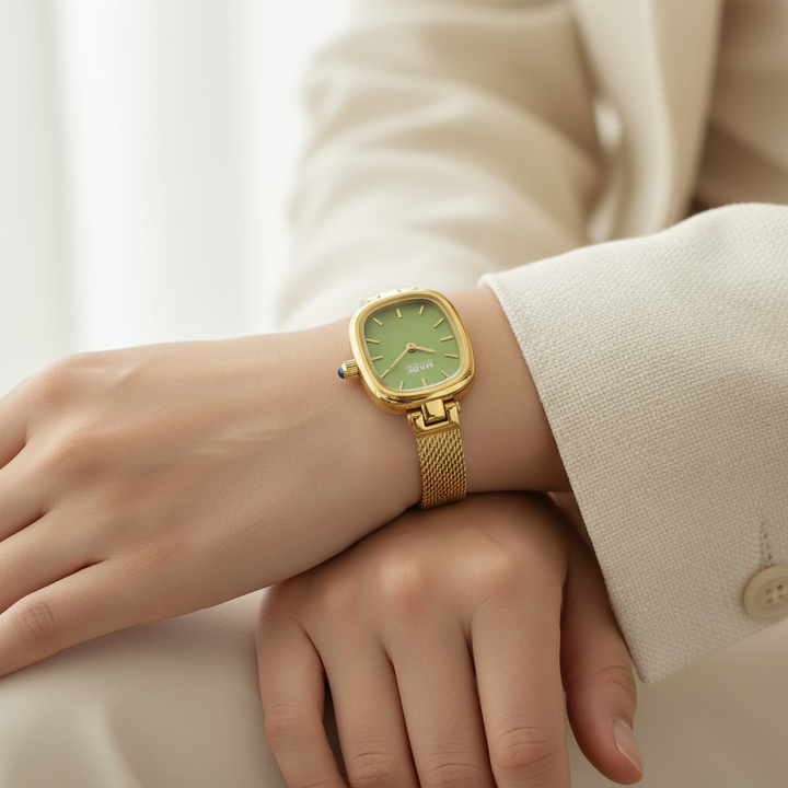 Gold watch with a green face on a wrist, wearing a beige outfit.