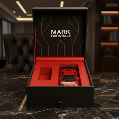 Red toy car in a black and red box with 'Mark Fairwhale' branding on a marble surface.