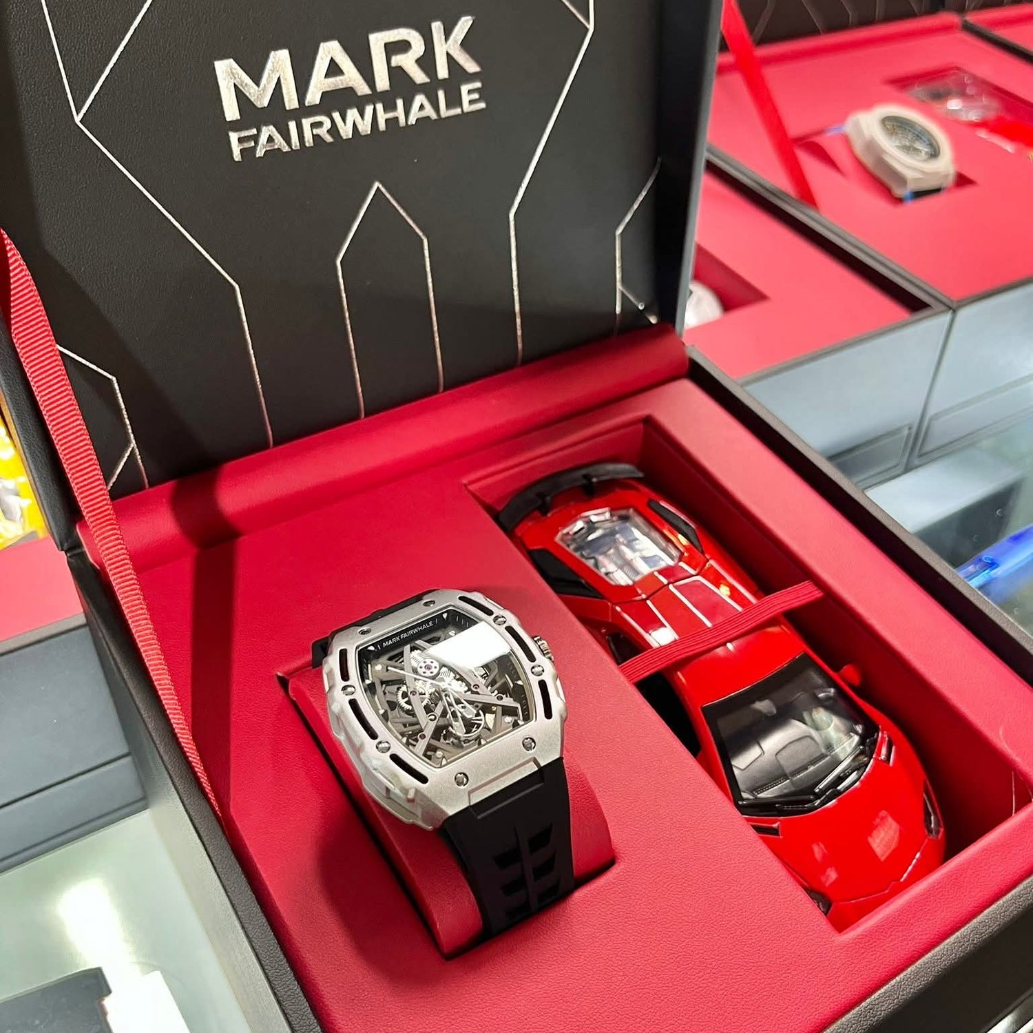 Mark Fairwhale luxury skeleton watch in silver case with black rubber strap presented in red premium packaging box next to miniature red sports car