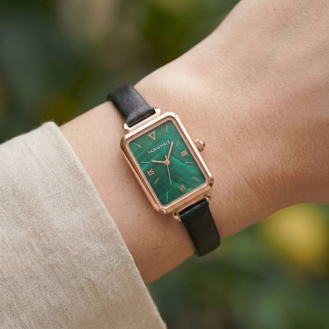 Wrist wearing a watch with a green face and black strap against a blurred natural background