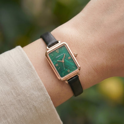 Wrist wearing a watch with a green face and black strap against a blurred natural background